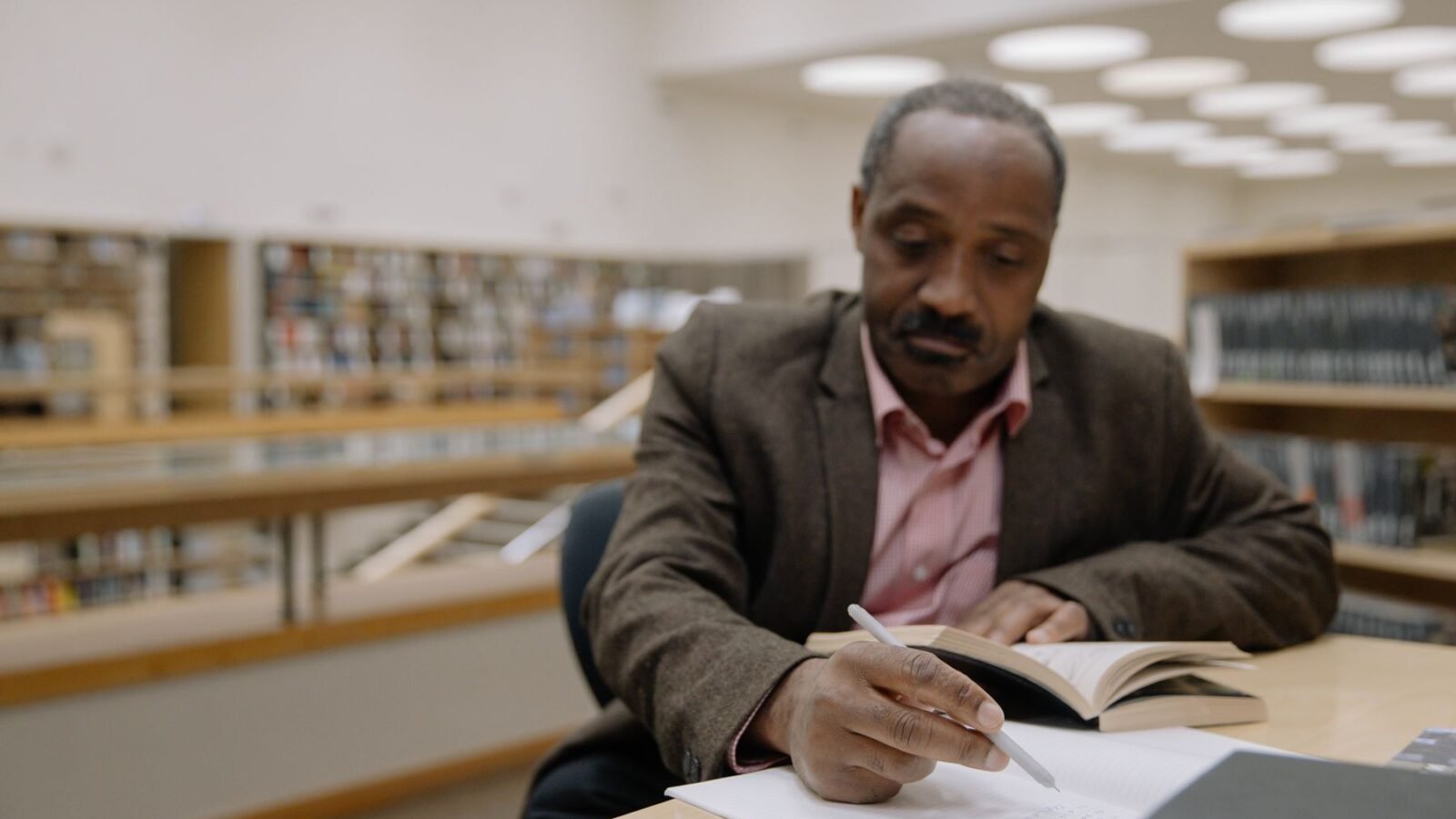 A man in a jacket writing notes at a desk in a library